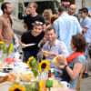 Das Bild zeigt eine Gruppe Menschen, die um einen gedeckten Tisch sitzen oder stehen. Auf dem Tisch sind Sonneblumen, Getränke und verschiedenes Essen zu sehen. Zwei Personen lächeln in die Kamera und zeigen ein 'Daumen hoch'