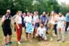 Das Bild zeigt eine Gruppe Menschen im Park. Es ist Sommer und die Bäume im Hintergrund tragen Blätter. Die Gruppe lacht und unterhält sich. Eine Person trägt ein Plakat von Start with a Friend. Darauf steht: Miteinander Füreinander.