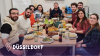 Das Foto zeigt eine Gruppe von 9 Menschen die gemeinsam Iftar in Düsseldorf feiern. Vor Ihnen ist ein Tisch voller verschiedener Gerichten zu sehen. Am unteren Rand ist ein dunkelblauer Farbverlauf zu sehen. Links unten ist eine Pinnadel und der Text 'Düsseldorf' in weiß zu sehen.