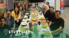 Das Foto zeigt eine Gruppe von 12 Menschen die gemeinsam Iftar in Stuttgart feiern. Jeweils 6 von Ihnen sitzen sich an einer langen Tafel gegenüber. Vor ihnen stehen Teller und Getränke. Sie lächeln in die Kamera. Am unteren Rand ist ein grüner Farbverlauf zu sehen. Links unten ist eine Pinnadel und der Text 'Stuttgart' in weiß zu sehen.